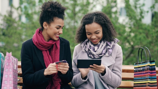 Duas mulheres com sacos de papel sentados no banco e usando computador tablet e cartão de crédito para fazer compras