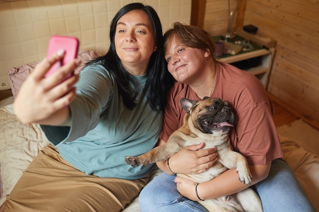 Duas mulheres com sobrepeso sentadas na cama e fazendo selfie no celular com seu animal de estimação