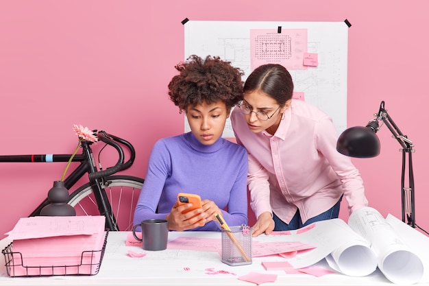 Foto duas mulheres criativas colegas de trabalho concentradas na tela do smartphone estudam as informações necessárias para fazer a pose do projeto na área de trabalho com papéis ao redor têm expressões sérias. pessoas trabalham em colaboração