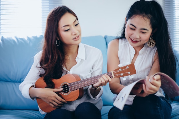 Duas mulheres da Ásia estão se divertindo tocando ukulele e sorrindo em casa para relaxar o tempo