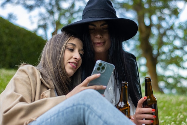 Duas mulheres de chapéu sentadas na grama brindando com garrafas de cerveja enquanto verificam seus telefones celulares Duas mulheres jovens de chapéu sentadas na grama bebendo vinho e usando seus telefones celulares