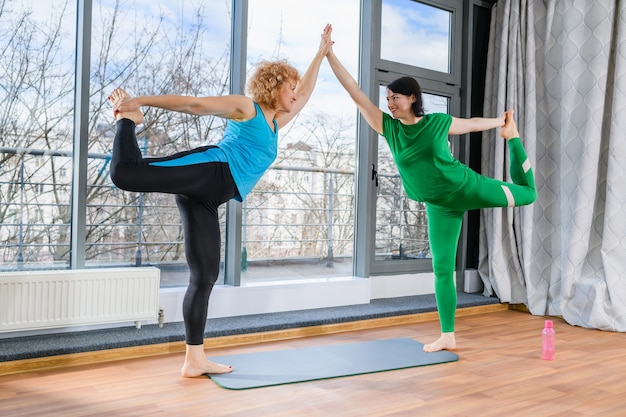 Duas mulheres de meia-idade se alongam e se equilibram juntas, de mãos dadas e apoiadas em uma perna só