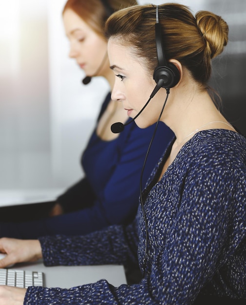 Duas mulheres de negócios conversam com os clientes por fones de ouvido, enquanto estão sentadas à mesa em um escritório moderno e ensolarado. Grupo de pessoas diversas em um call center. Telemarketing e atendimento ao cliente.