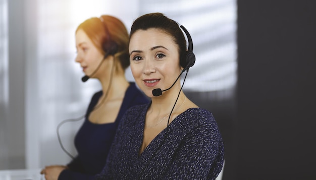 Duas mulheres de negócios conversam com os clientes por fones de ouvido, enquanto estão sentadas à mesa em um escritório moderno e ensolarado. Grupo de pessoas diversas em um call center. Telemarketing e atendimento ao cliente.