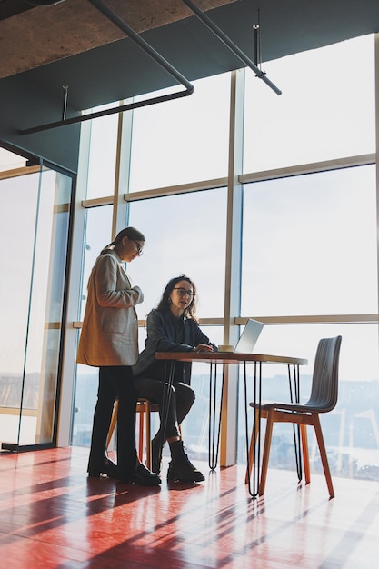 Duas mulheres de negócios estão procurando algo em um laptop Conceito de cooperação empresarial e trabalho em equipe Jovens mulheres sorridentes na mesa do escritório Pessoas modernas de sucesso