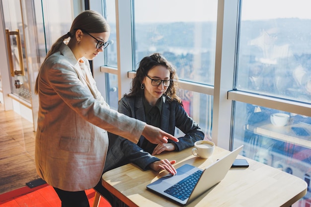 Duas mulheres de negócios estão procurando algo em um laptop Conceito de cooperação empresarial e trabalho em equipe Jovens mulheres sorridentes na mesa do escritório Pessoas modernas de sucesso