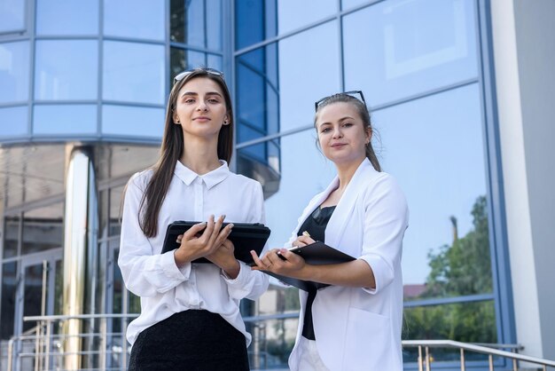 Duas mulheres de negócios jovens e bem-sucedidas com um tablet em pé perto de um prédio comercial