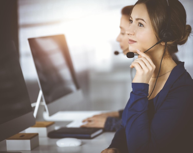 Duas mulheres de negócios têm conversas com os clientes por fones de ouvido enquanto estão sentadas na mesa em um escritório moderno ensolarado Grupo de pessoas diversas em um call center Telemarketing e atendimento ao cliente