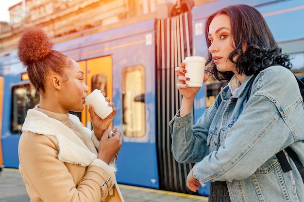 Duas mulheres em jaquetas jeans conversando rindo bebendo café e esperando o bonde