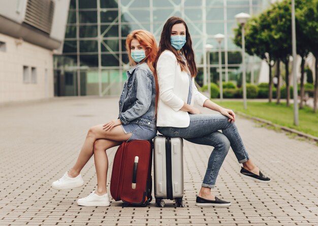 Duas mulheres em máscaras protetoras, sentado em malas, esperando o voo