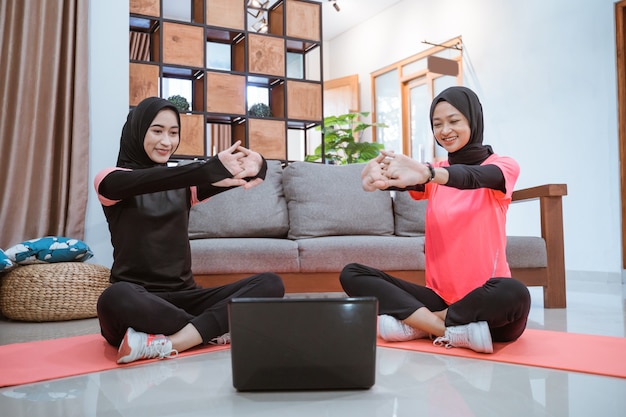 Duas mulheres em roupas esportivas hijab sentadas no chão, aquecendo, esticando os braços para a frente, enquanto fazem atividades juntas em casa