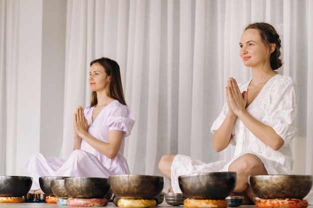 Duas mulheres estão sentadas com tigelas tibetanas na posição de lótus antes de uma aula de ioga no ginásio