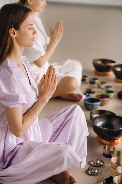 Duas mulheres estão sentadas com tigelas tibetanas na posição de lótus antes de uma aula de ioga no ginásio