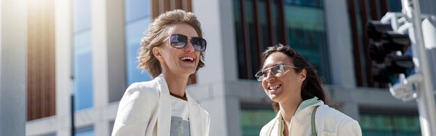 Duas mulheres felizes em roupas da moda riem e caminham para fora no fundo de um edifício moderno