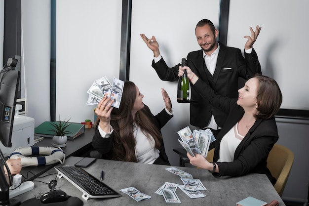 Duas mulheres felizes sentadas na mesa segurando um pacote de dinheiro e uma garrafa de vinho comemorando o grande evento Colegas animados felizes comemorando o triunfo juntos no escritório com champanhe Homem surpreso