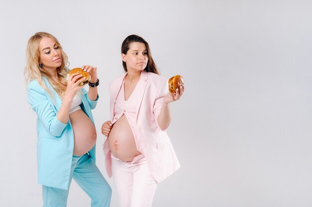 Duas mulheres grávidas de terno com hambúrgueres nas mãos em um fundo cinza