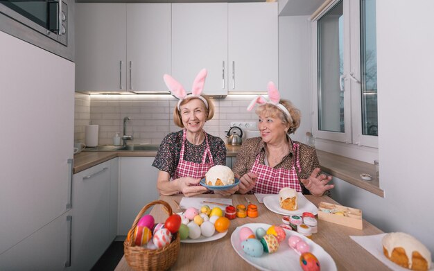 Duas mulheres idosas com orelhas de coelho na cabeça estão pintando ovos de Páscoa em casa na cozinha