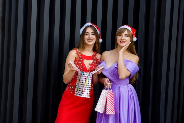 Duas mulheres incríveis com chapéu de Papai Noel e vestidos de noite elegantes segurando sacolas de compras