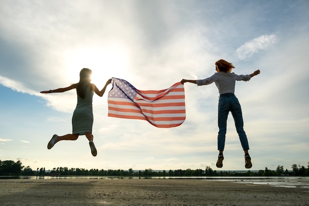 Duas mulheres jovens amigos segurando a bandeira nacional dos EUA, pulando juntos ao ar livre ao pôr do sol. Silhueta de meninas comemorando o dia da independência dos Estados Unidos. Dia internacional do conceito de democracia.