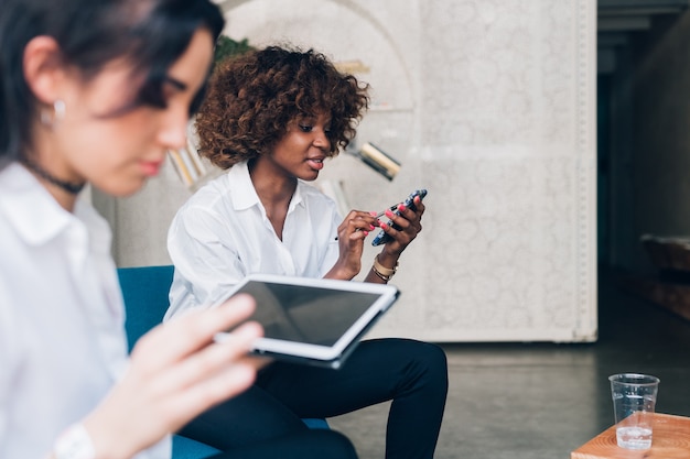 Duas mulheres jovens multirraciais, trabalhando com dispositivo digital no escritório moderno