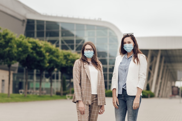 Duas mulheres jovens no fundo de um prédio ao ar livre, com máscaras médicas no rosto