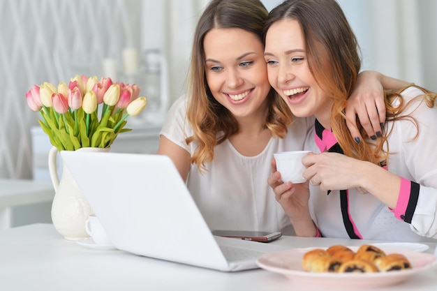 Duas mulheres jovens olhando para laptop enquanto está sentado na mesa da cozinha com biscoitos