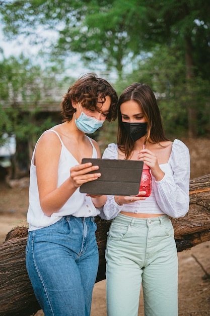 Duas mulheres jovens olhando para um computador tabletEles estão usando máscaras médicas e se apoiando em uma árvore