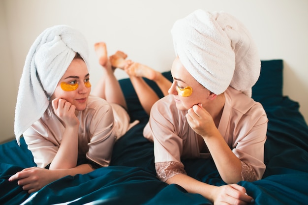 Duas mulheres jovens se divertindo com manchas sob os olhos. Dois amigos de toalhas e pijamas fazem uma divertida festa no spa juntos em casa.