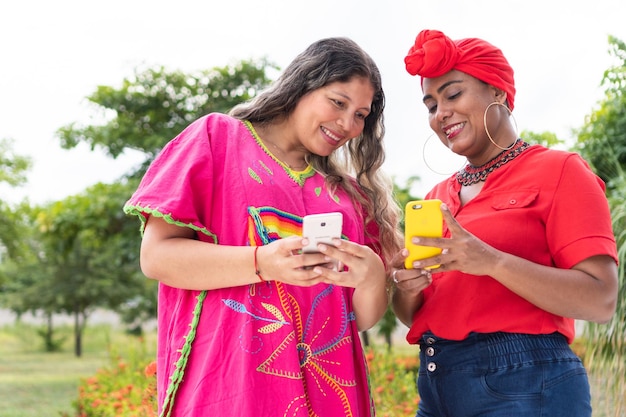 Duas mulheres latinas em pé no parque olhando para o celular e conversando