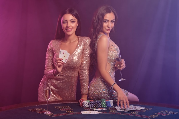Duas mulheres lindas mostrando cartas enquanto posam na mesa de jogo no cassino Fundo de fumaça preta com luzes coloridas Jogo de pôquer Closeup
