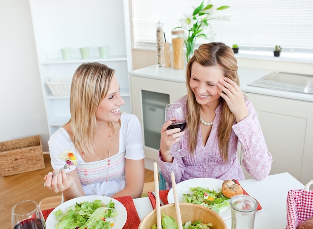 Duas mulheres na cozinha conversando e comendo salada com copos de vinho tinto