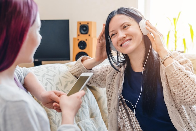 Duas mulheres ouvindo música no smartphone com fones de ouvido sentado no sofá em casa