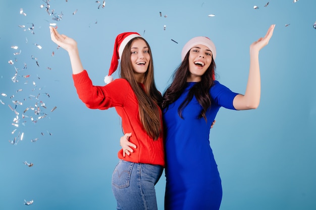 Duas mulheres sorridentes usando chapéus de Papai Noel jogam confete prata no ar isolado sobre o azul