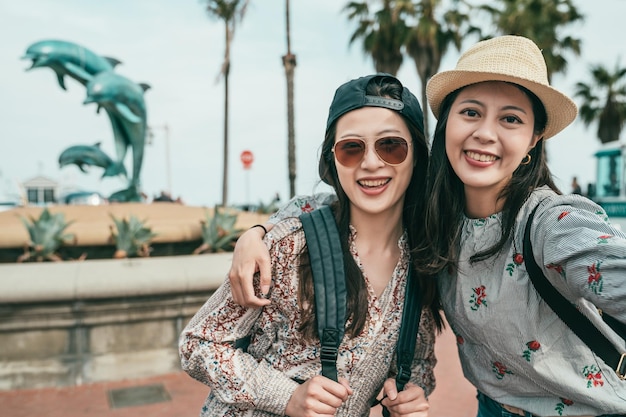 duas mulheres sorrindo e tirando selfies alegremente em frente a uma fonte de golfinhos em uma linda praça.