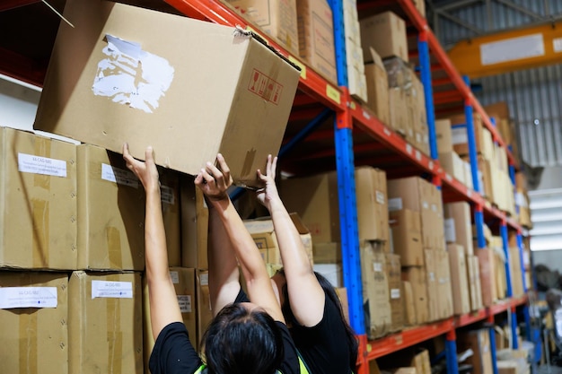 Foto duas mulheres trabalhadoras carregando e levantando caixa de papelão pesada trabalhadora de armazém obesa asiática em colete de segurança e capacete de capacete trabalhando em fábrica de armazém industrial