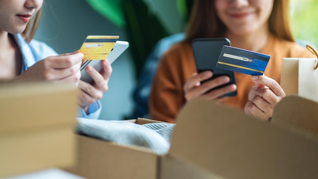 Duas mulheres usando telefone celular e cartão de crédito para compras on-line com sacola de compras e caixa postal de roupas em cima da mesa