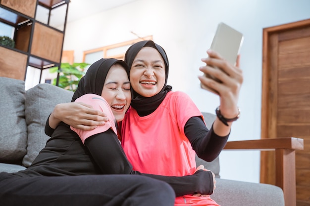Duas mulheres vestindo roupas esportivas hijab riem e se abraçam durante uma videochamada com um celular, sentadas no chão da casa