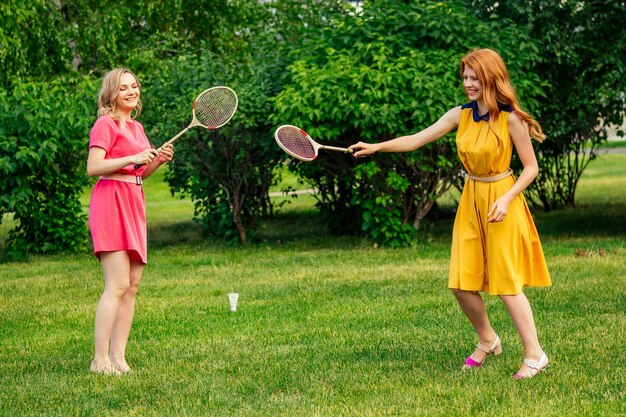 Duas namoradas engraçadas linda jovem ruiva irlandesa ruiva em um vestido amarelo e uma mulher loira europeia em um vestido rosa jogando tênis com raquete de badminton no parque de verão