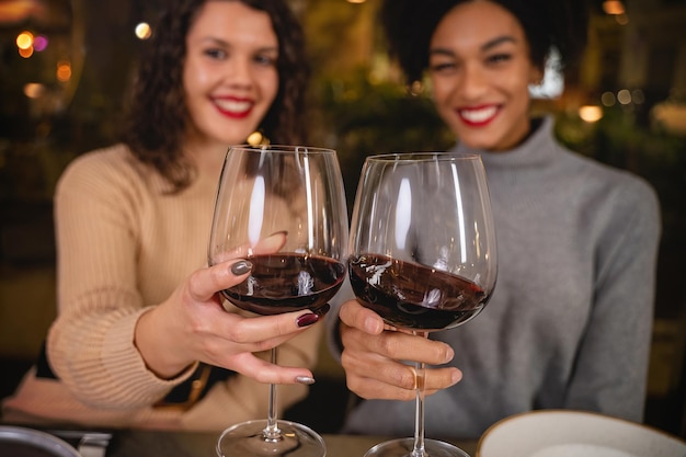 Duas namoradas multiculturais comemorando um brinde de vinho tinto em um bar restaurante, olhando para a câmera - conceito de pessoas bebendo álcool na festa