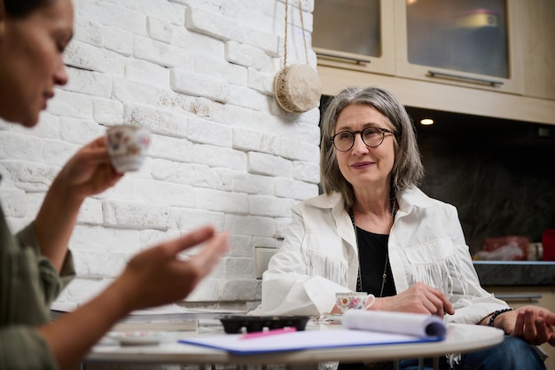 Duas pessoas diversas de duas gerações sentadas em uma mesa e desfrutando de uma discussão enquanto bebem café em um estúdio de design de moda aconchegante