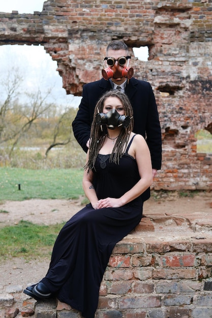 Duas pessoas em roupas pretas e máscaras de gás posam contra o fundo de uma parede de tijolos destruída