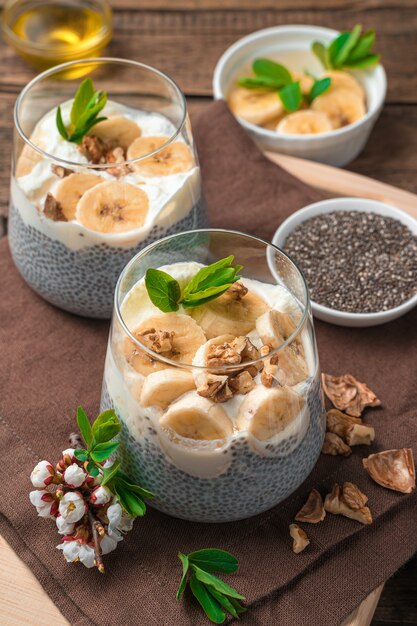 Duas porções de sobremesa natural saudável com sementes de chia em leite vegetal com banana, nozes e mel em uma parede de madeira marrom. Vista lateral, vertical.