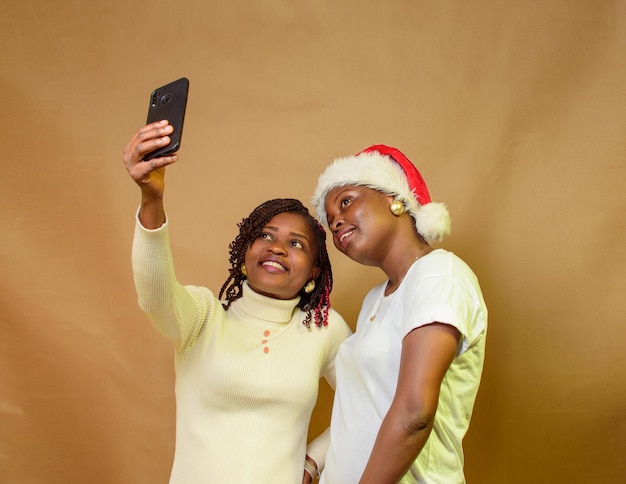 Foto duas senhoras, irmãs ou amigas africanas tirando fotos com um smartphone enquanto uma delas tem um boné de natal na cabeça