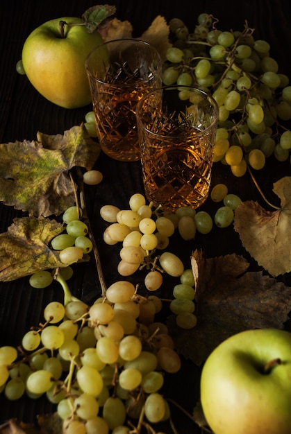 Duas taças de cristal com vinho branco, maçãs, uvas e folhas secas em uma superfície de madeira