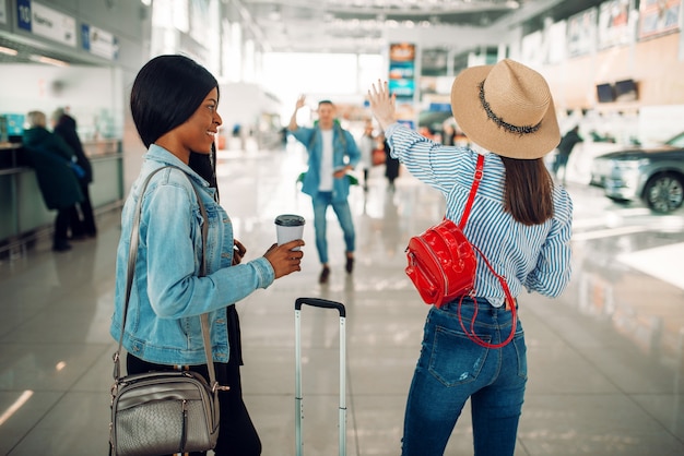 Duas turistas com casos encontram um amigo no aeroporto. Passageiros com bagagem no terminal aéreo