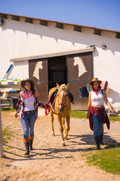 Duas vaqueiras saindo sorrindo puxando um cavalo para fora de um estábulo, com roupas sul-americanas, estilo de vida de duas trabalhando em um estábulo