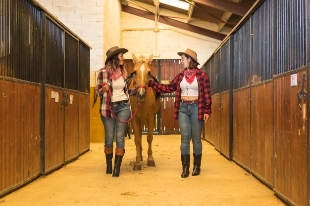 Duas vaqueiras se divertindo e rindo com um cavalo em um estábulo, chapéus de cowboy americanos, camisas xadrez e jeans