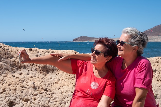 Duas velhas amigas sentadas em um banco olham para o celular e tiram uma selfie. Conceito de vínculo e amizade. Montanha e mar atrás deles. Paraíso para os amantes do surf