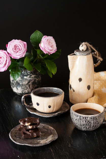 Duas xícaras de café preto, chocolate preto, um bule de chá amarelo e um vaso com rosas em uma superfície escura