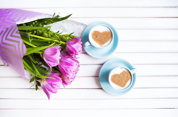 Duas xícaras de cappuccino com um símbolo em forma de coração e tulipas roxas em um fundo de madeira